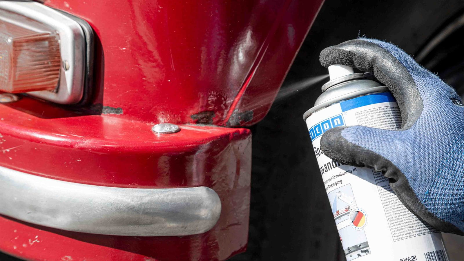 Rostumwandler wird aus der Spraydose auf eine angerostete Stelle an einem roten Fahrzeugblech gesprüht